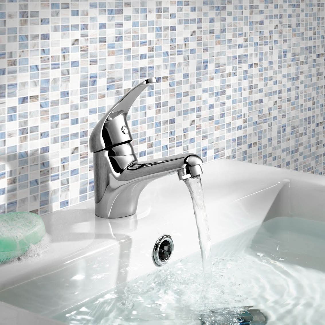 The iBathUK Orvex Monobloc Basin Mixer Tap runs water into a white basin, partially filled. A green soap bar is nearby, with blue and white mosaic tiles on the backsplash adding a modern touch.