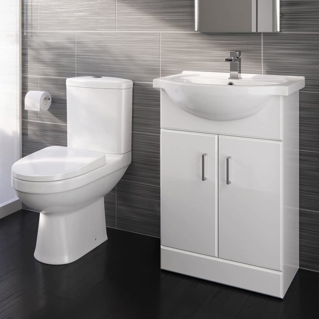 A modern bathroom features the iBathUK Bathroom Ceramic Suite: a vanity sink unit with soft-close doors and a close-coupled toilet, both in white, set against gray tiled walls and a dark floor with a wall-mounted toilet paper holder.