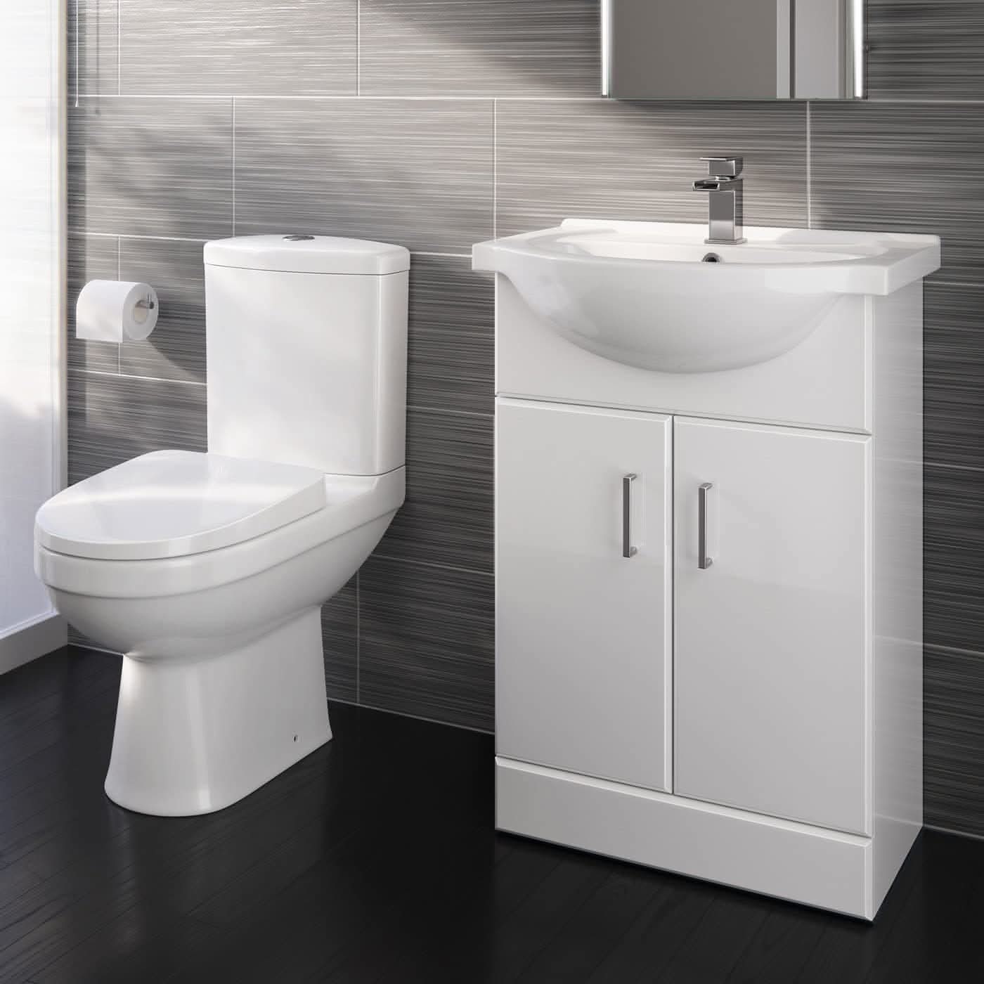 A modern bathroom features the iBathUK Bathroom Ceramic Suite: a vanity sink unit with soft-close doors and a close-coupled toilet, both in white, set against gray tiled walls and a dark floor with a wall-mounted toilet paper holder.