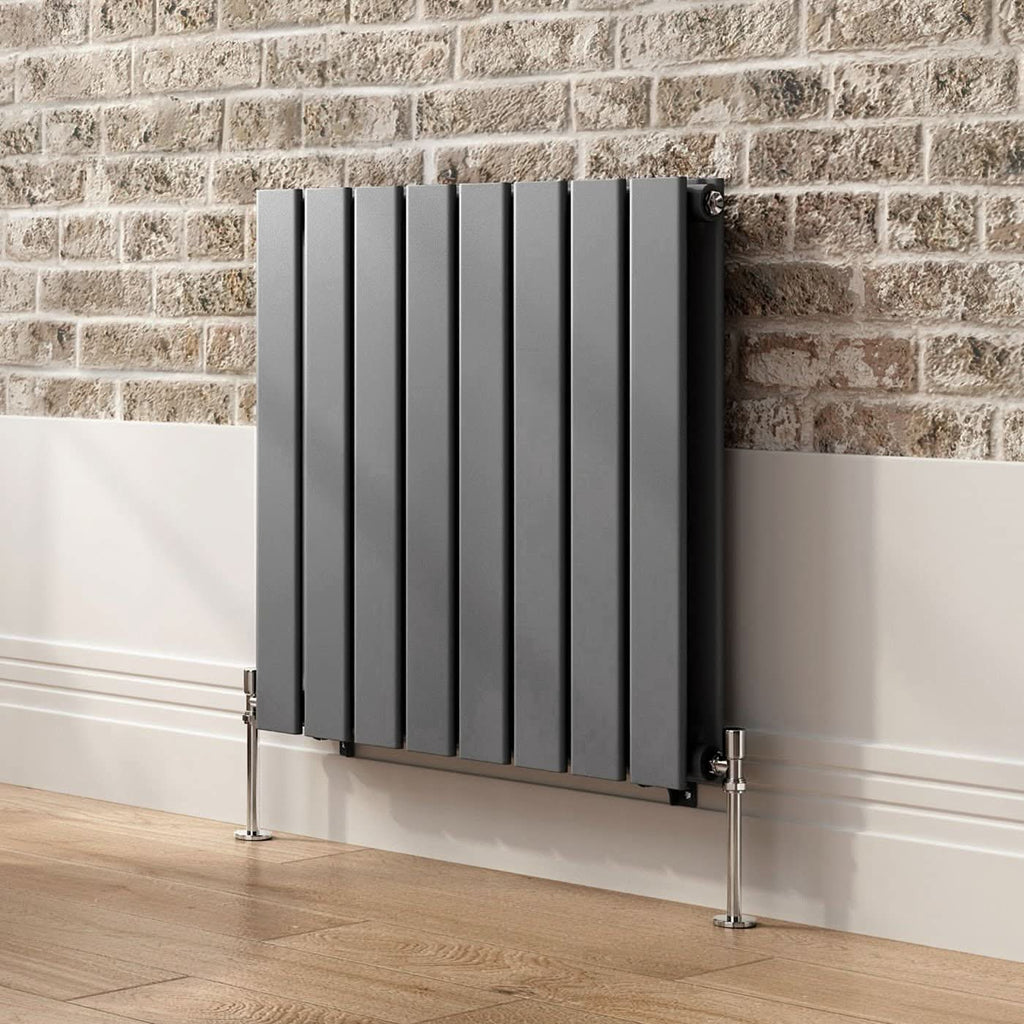 The iBathUK Apex Horizontal Double Slim Flat Panel Designer Radiator is mounted on a white wall with a brick accent, standing on light wood flooring with visible chrome pipes.