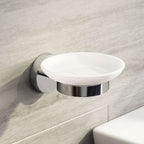 The iBathUK Lumo Designer Wall Mounted Soap Dish, part of a chrome accessory set with toothbrush holder and tumbler, features a white round tray and chrome base fixed above a white sink on a light gray tiled bathroom wall.