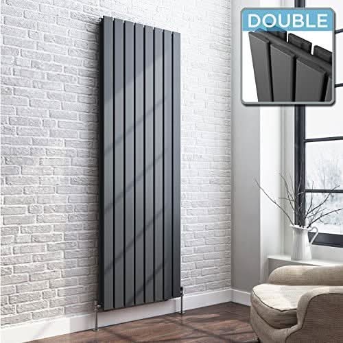 The iBathUK Apex Vertical Double Panel Slim Flat Designer Radiator in matt anthracite is mounted on a white brick wall in a modern room with a window, beige chair, and small vase. An inset highlights the radiator’s sleek panels.
