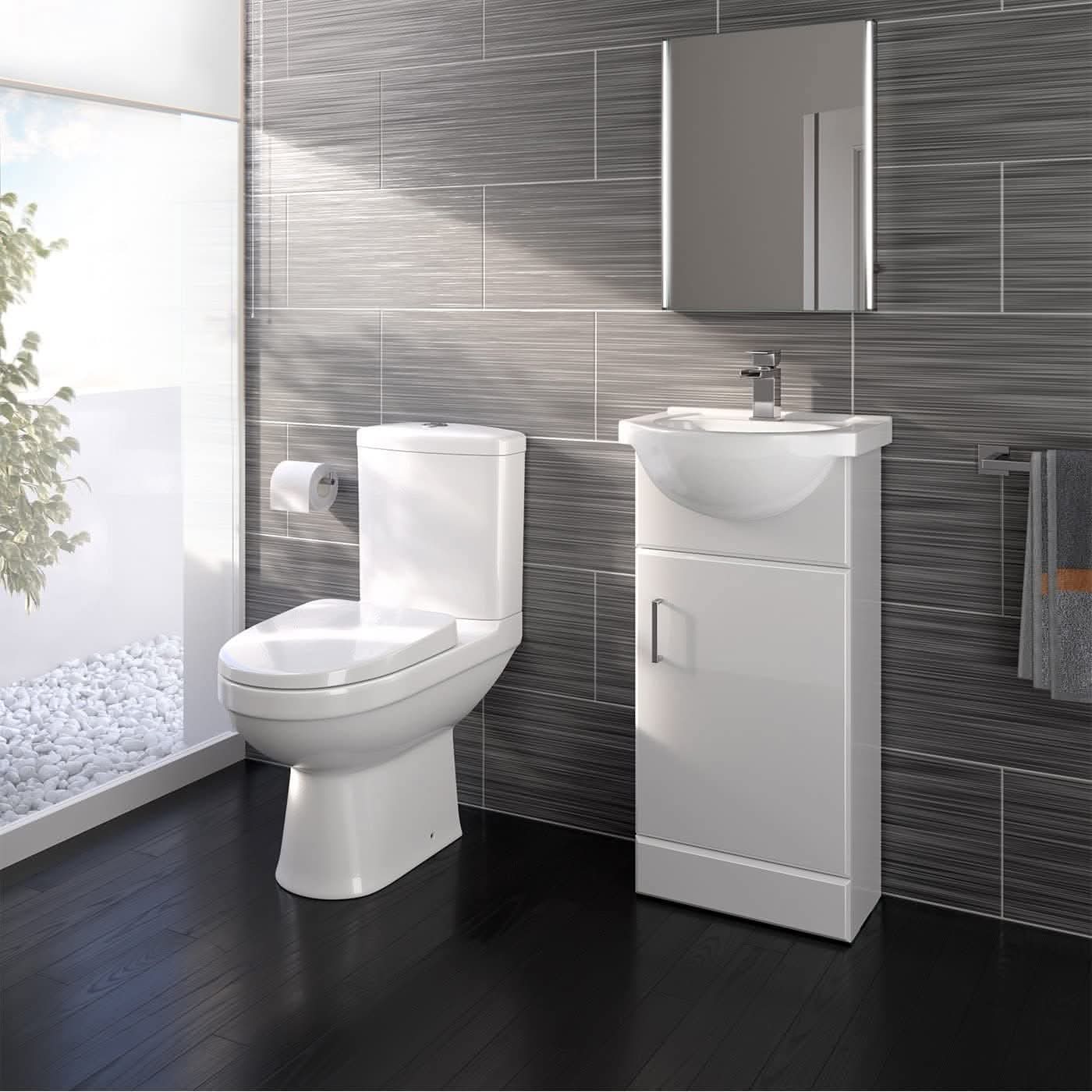 The iBathUK Petite Modern Bathroom Suite - Gloss White features a white toilet, sink cabinet, rectangular mirror, and towel rail on gray tiles. A large frosted window shows white pebbles and greenery; the dark wood floor enhances elegance.