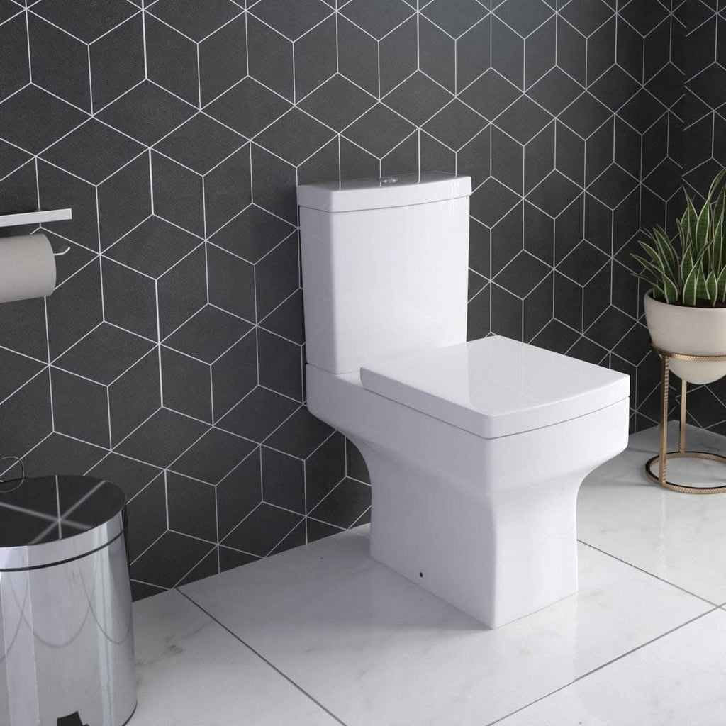 An iBathUK Avon Modern Bathroom Suite in gloss white features a modern toilet set against geometric black and white tiles, with a chrome trash can, toilet paper holder, and a potted plant on a spotless white floor.