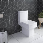An iBathUK Avon Modern Bathroom Suite in gloss white features a modern toilet set against geometric black and white tiles, with a chrome trash can, toilet paper holder, and a potted plant on a spotless white floor.