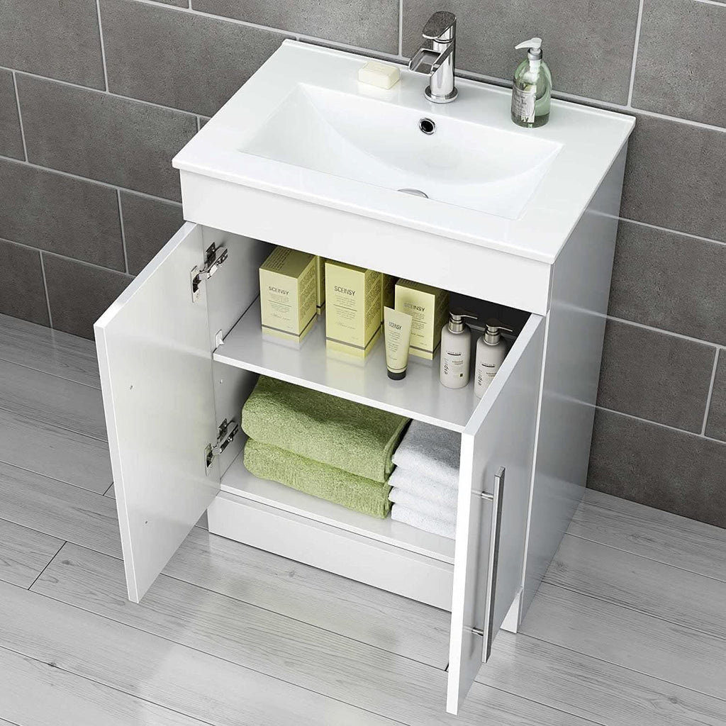 The iBathUK Avon Modern Bathroom Suite in Gloss White includes an open cabinet with green and white towels, boxed skincare products, and bottles beneath a white sink, all set against gray tiled walls and light wood flooring.