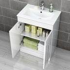 The iBathUK Avon Modern Bathroom Suite in Gloss White includes an open cabinet with green and white towels, boxed skincare products, and bottles beneath a white sink, all set against gray tiled walls and light wood flooring.