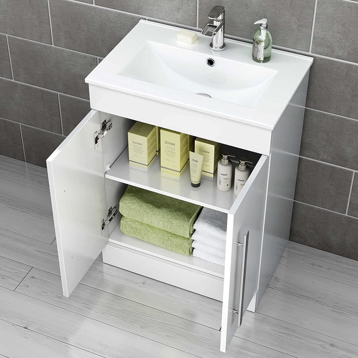 The iBathUK Avon 2 Door Floorstanding Vanity Unit With Basin (615mm x 815mm, 1 Tap Hole) stands against gray tiled walls and light wood flooring, its open cabinet showcasing neatly stacked towels and assorted toiletries.