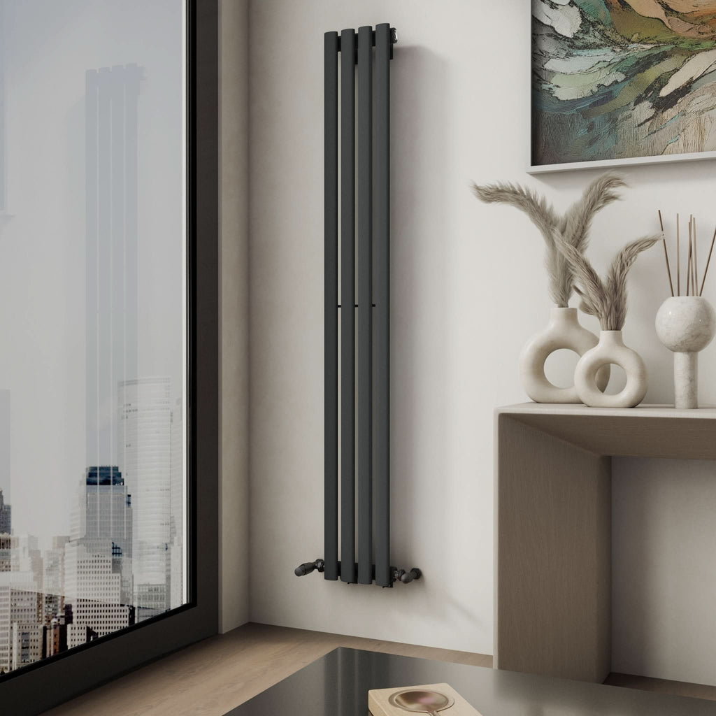 A modern room corner features the iBathUK Princia Vertical Single Oval Panel Radiator on a white wall beside a city-view window. A table displays vases with dried grasses, and an abstract painting above adds a stylish accent.