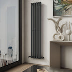 A modern room corner showcases the iBathUK Princia Vertical Single Oval Panel Radiator mounted on the wall near a large window, with a chic console table nearby holding vases, dried grasses, and abstract wall art above.