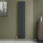 A Princia Vertical Single Oval Panel Radiator by iBathUK in black is mounted on an olive green panelled wall, accented by modern decor like a painting, vase, and dark cabinet to emphasize the radiators sleek, minimalist style.