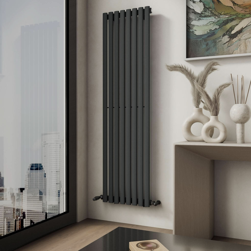 The Princia Vertical Single Oval Panel Radiator by iBathUK is mounted on the wall near a large window overlooking the city. A console table with decorative vases and pampas grass sits nearby beneath a vibrant abstract painting.