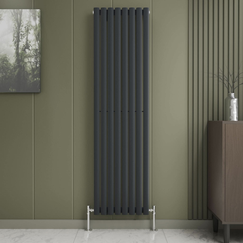 The iBathUK Princia Vertical Single Oval Panel Radiator is mounted on an olive-green wall beside decorative panels. A small cabinet with a vase stands to the right, with a framed nature print to the left above white tile flooring.