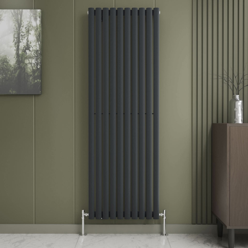 The iBathUK Princia Vertical Single Oval Panel Radiator is mounted on an olive-green wall with minimalist decor, including a small abstract painting, a vase with twigs on a brown cabinet, and a light marble floor.
