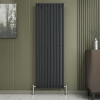 The iBathUK Princia Vertical Single Oval Panel Radiator is mounted on an olive-green wall with minimalist decor, including a small abstract painting, a vase with twigs on a brown cabinet, and a light marble floor.
