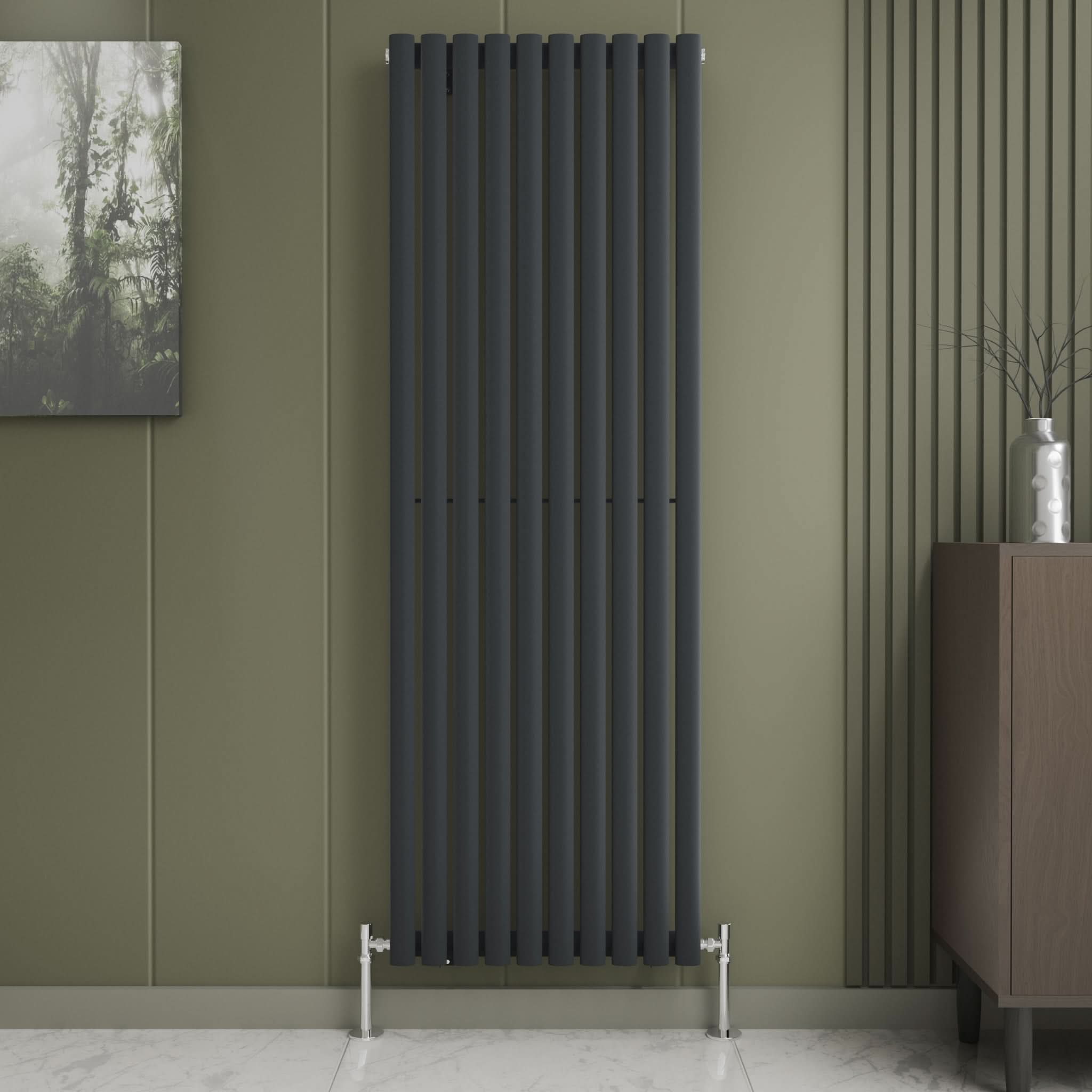 The iBathUK Princia Vertical Single Oval Panel Radiator is mounted on an olive-green wall with minimalist decor, including a small abstract painting, a vase with twigs on a brown cabinet, and a light marble floor.