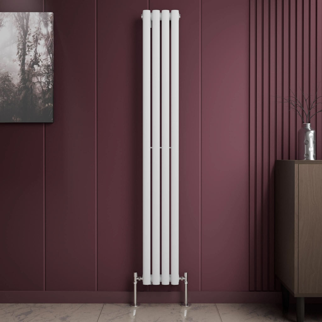 The iBathUK Princia Vertical Single Oval Panel Radiator in white stands against a dark mauve wall with decorative grooves, flanked by a framed picture on the left and a cabinet with a vase and branches on the right, atop light-colored tile flooring.