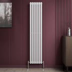 The iBathUK Princia Vertical Single Oval Panel Radiator in white stands against a burgundy panelled wall beside a brown cabinet and wall art. The light-toned floor features a subtle marble pattern.