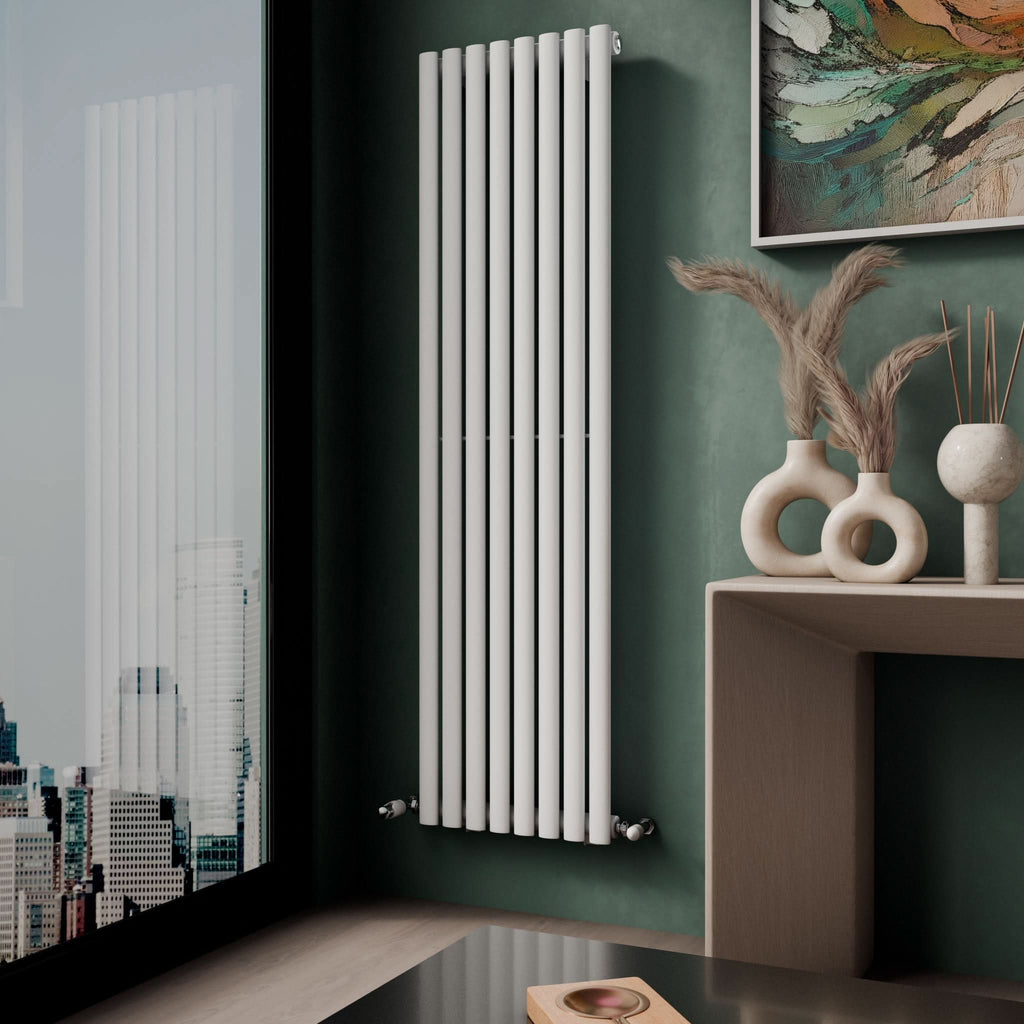 A modern room features the iBathUK Princia Vertical Single Oval Panel Radiator on a green wall, a wooden shelf with decorative vases and dried grasses, and a large window overlooking the city.