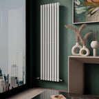 A modern room features the iBathUK Princia Vertical Single Oval Panel Radiator on a green wall, a wooden shelf with decorative vases and dried grasses, and a large window overlooking the city.