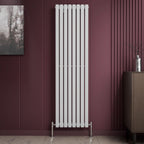 A Princia Vertical Single Oval Panel Radiator by iBathUK, in white, is mounted on a maroon wall. The room has a small cabinet with a silver vase, framed picture, and a light-colored floor.