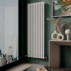 A modern room showcases the iBathUK Princia Vertical Single Oval Panel Radiator on a green wall near a window with city views. A beige table displays vases of dried plants and a round sculpture, with an abstract painting above.