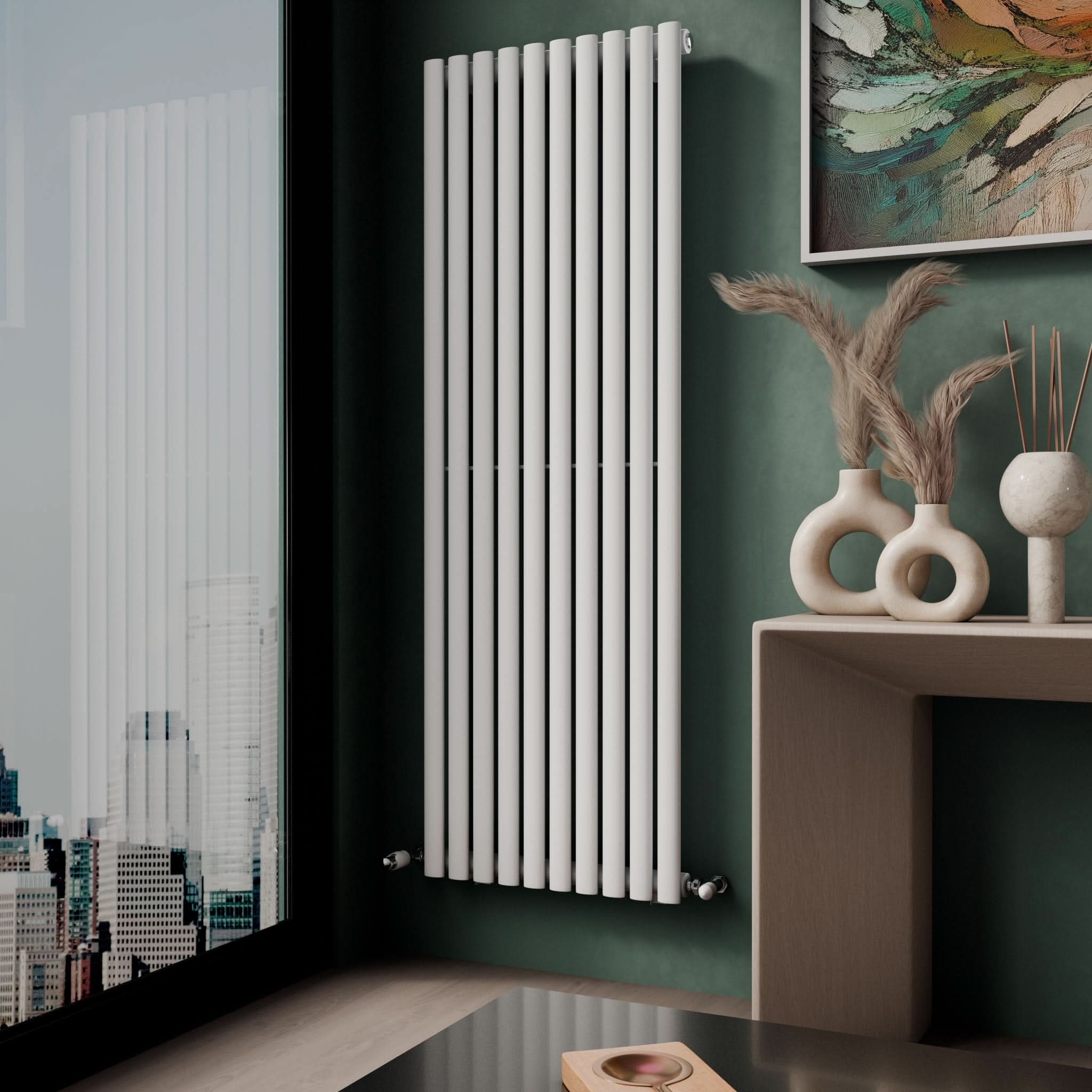 A modern room showcases the iBathUK Princia Vertical Single Oval Panel Radiator on a green wall near a window with city views. A beige table displays vases of dried plants and a round sculpture, with an abstract painting above.